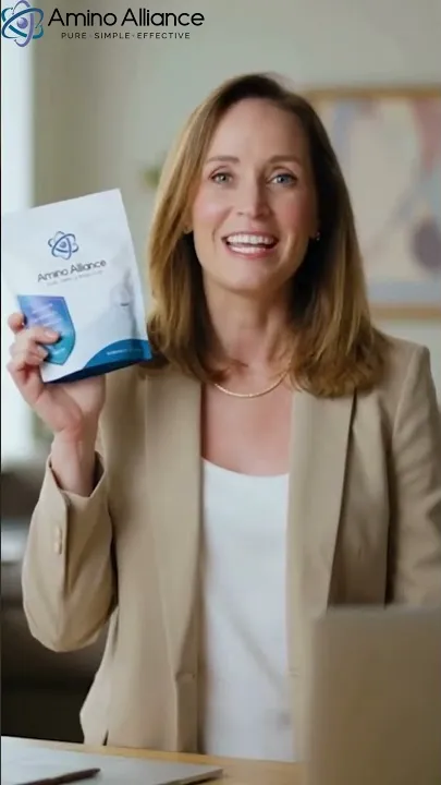 A woman in a tan blazer over a white top holding an Amino Alliance pouch up to camera in a bright office, a laptop at the edge of frame and framed artwork behind her.