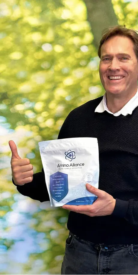 A man in a black jumper holding an Amino Alliance pouch to camera with a thumbs up, green foliage behind him.