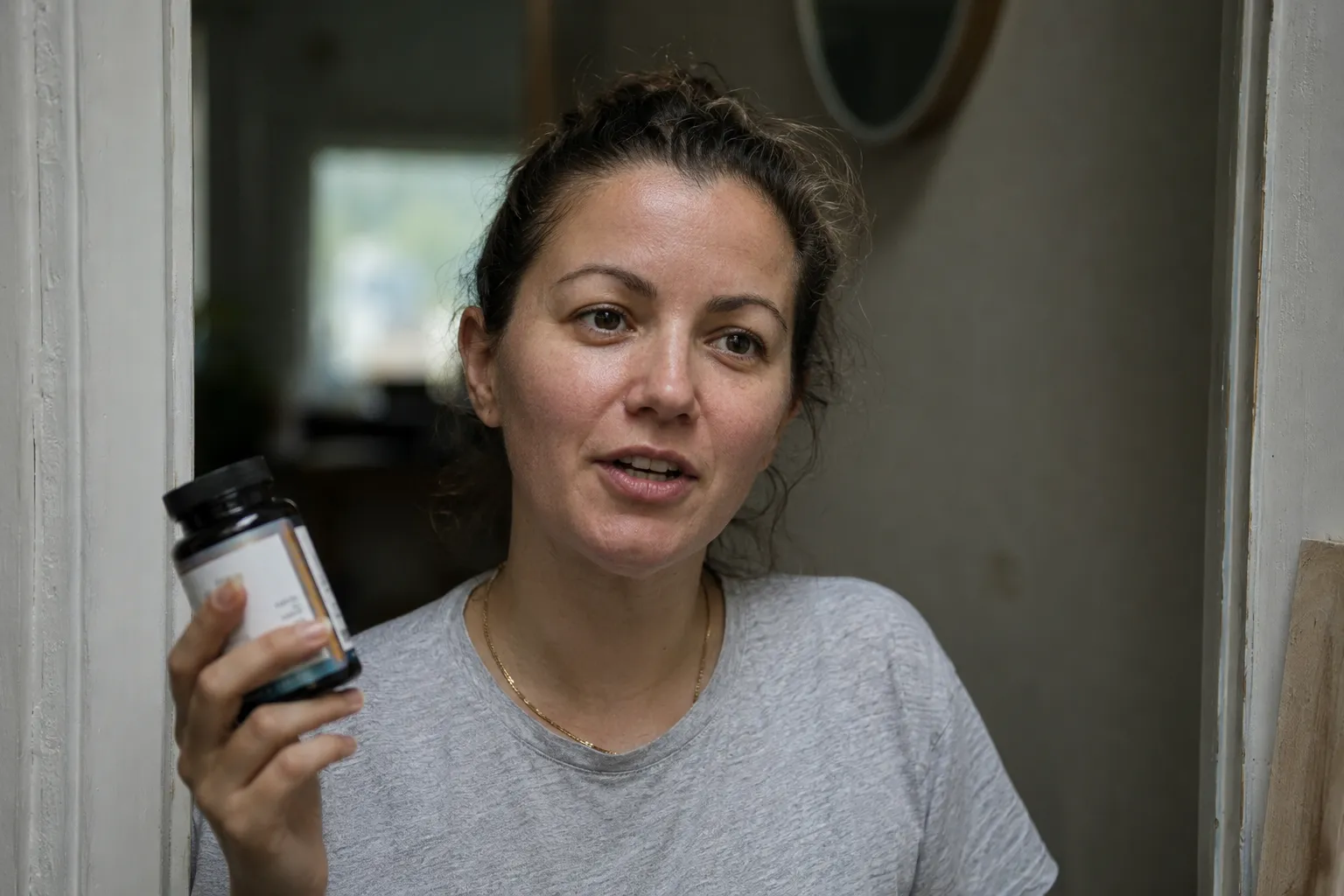 A woman in a grey t-shirt stands in a doorway holding up a dark supplement jar toward the camera, mid-sentence, with an unmade room and a mirror behind her.