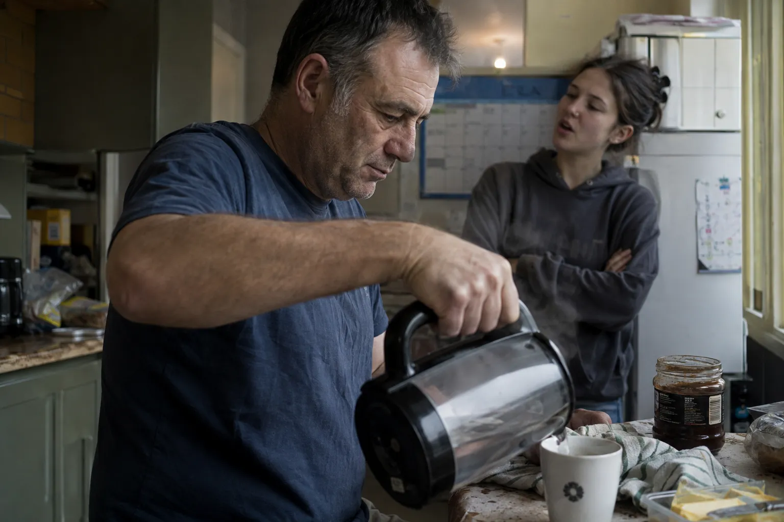 A man in a navy t-shirt pours from a glass coffee jug into a mug in a cluttered domestic kitchen while a young woman in a grey hoodie stands behind him with her arms folded, mid-sentence, looking at him.