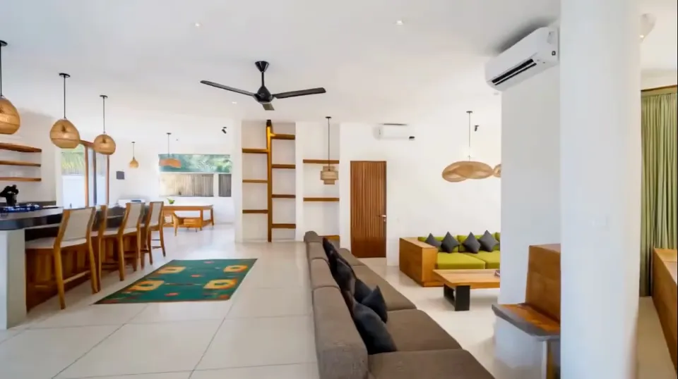 The open-plan living space of a Bali villa: polished floor, a low sofa under a ceiling fan, pendant lights over a kitchen counter and a green rug down the middle.