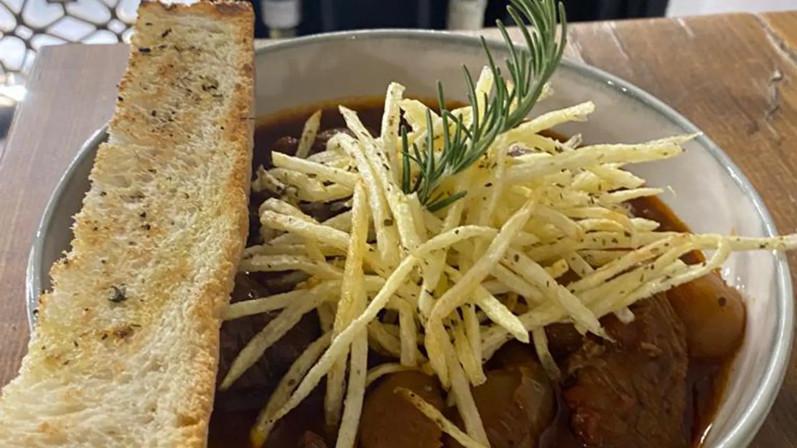 Beef stifado in a dark sauce in a metal bowl under a nest of shoestring fries and a sprig of rosemary.