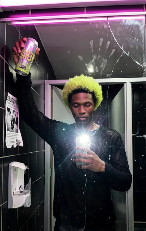 The same man holding a FERAL can up in a bathroom mirror selfie, flash on, pink strip light above the cracked mirror.