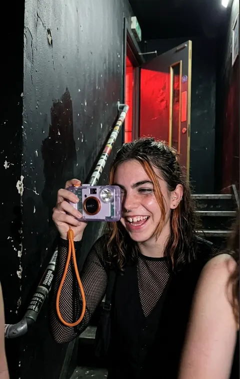 The same woman laughing in a dark club stairwell, raising the compact camera to her eye, red light behind her.