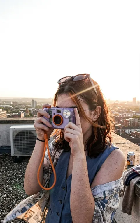 The same woman on a rooftop at sunset, camera to her eye, city and air conditioning units behind her.