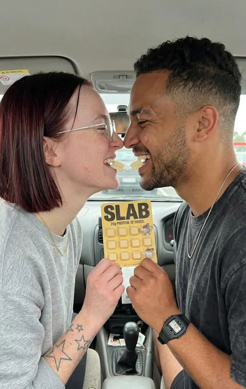 Two people in a car holding a SLAB tray between them, both mid-laugh, faces close together.